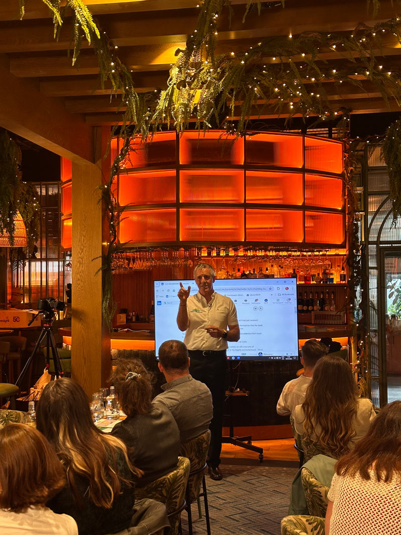 A group of people in a venue listening to a man present a talk. There is greenery on roof and orange lightning.