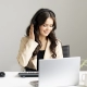 Call Agent smiling while on a call. She is sat infront of white desk wearing a black top and cream jacket. A laptop, keypad, mug and plant are on desk.