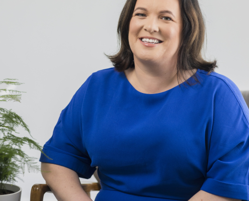 Woman smiling in blue dress sat in front of white wall with plant next to her.