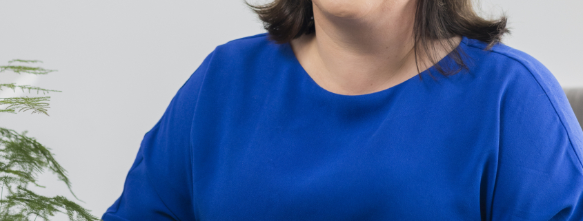 Woman smiling in blue dress sat in front of white wall with plant next to her.