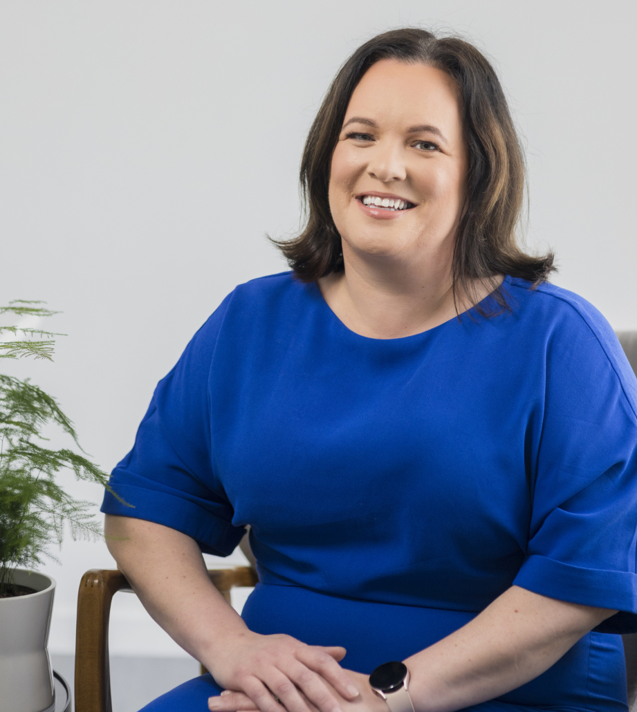 Woman smiling in blue dress sat in front of white wall with plant next to her.