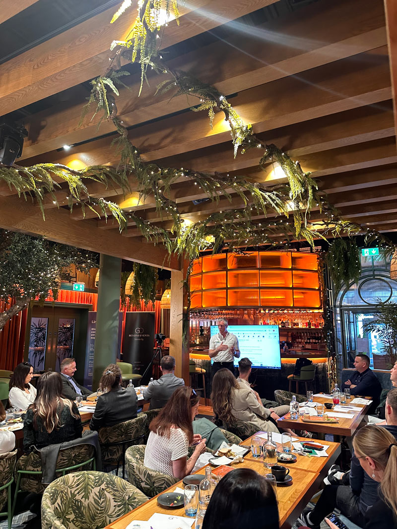 A group of people in a venue listening to a man present a talk. There is greenery on roof and orange lightning.