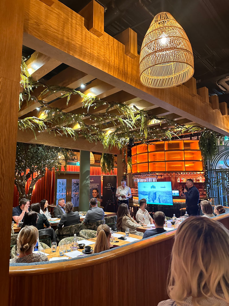 A group of people in a venue listening to a man present a talk. There is greenery on roof and orange lightning.