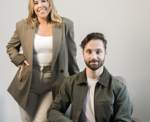 A call agent standing behind a sitting call agent. They are wearing green outfits with a white wall behind them.