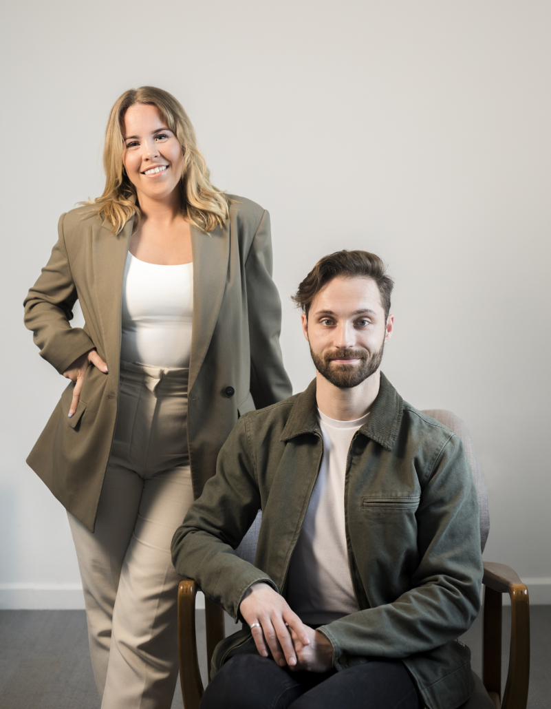 A call agent standing behind a sitting call agent. They are wearing green outfits with a white wall behind them.