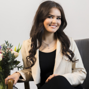 A woman with long brown hair is wearing a black top and trousers with a cream blazer. She is sat on a chair in front of a grey wall beside flowers.