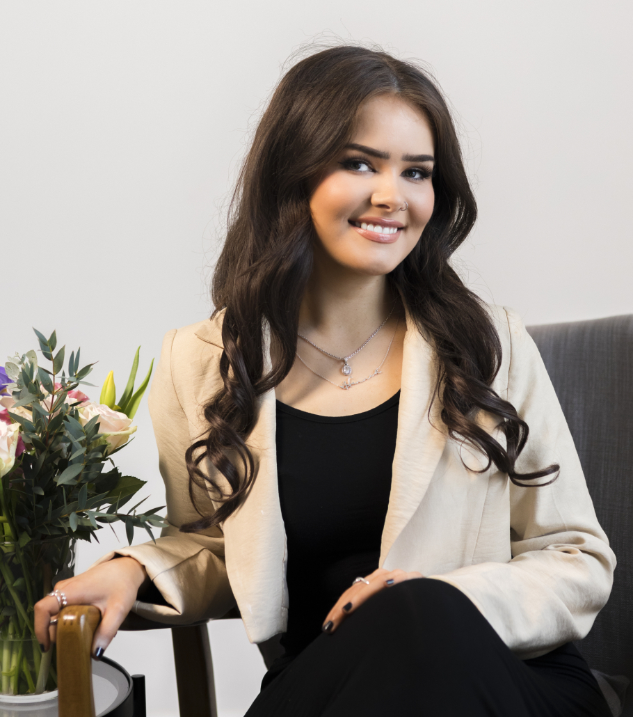 A woman with long brown hair is wearing a black top and trousers with a cream blazer. She is sat on a chair in front of a grey wall beside flowers.