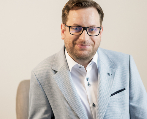 Man wearing white shirt and blue blazer sat infront of grey wall.