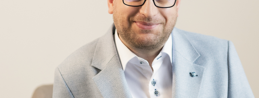 Man wearing white shirt and blue blazer sat infront of grey wall.