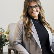 A woman wearing glasses sat in front of grey wall wearing a black top and grey blazer.