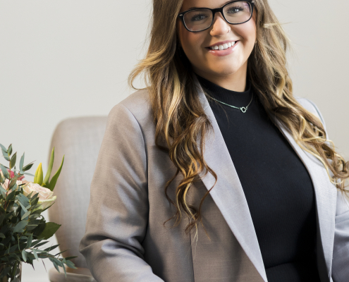 A woman wearing glasses sat in front of grey wall wearing a black top and grey blazer.