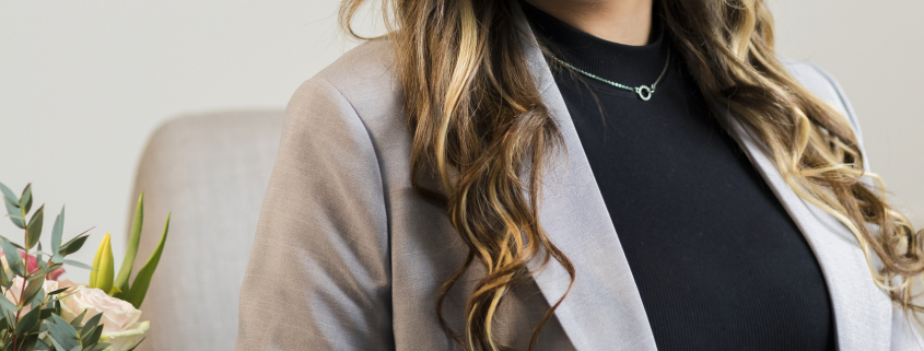 A woman wearing glasses sat in front of grey wall wearing a black top and grey blazer.