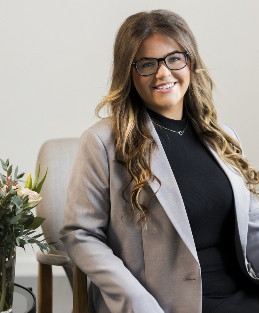 A woman wearing glasses sat in front of grey wall wearing a black top and grey blazer.